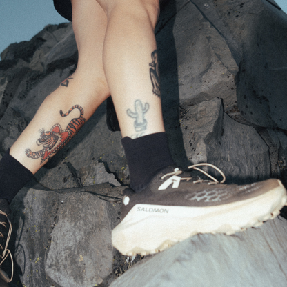 A close-up lifestyle shot of a person's legs and feet wearing Salomon trail running shoes and black socks while sitting on dark rocks outdoors.