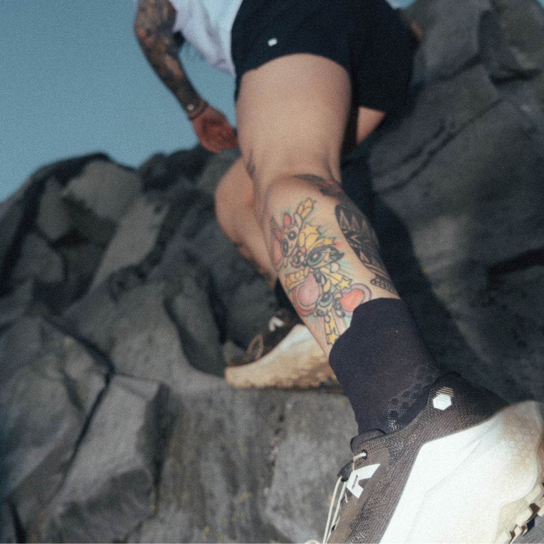 A close-up lifestyle shot of a person climbing rocky terrain, focusing on their tattooed lower leg wearing a black crew sock and a brown and white running shoe.