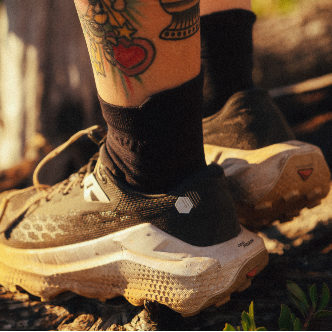 Close-up of a person wearing black athletic socks and trail running shoes outdoors on a rocky surface, with warm sunlight and visible tattoos on the lower leg.
