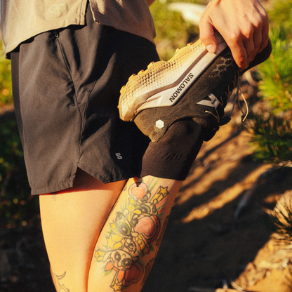 A close-up lifestyle shot of a person outdoors wearing black athletic shorts and socks, stretching their leg to put on a black Salomon running shoe.