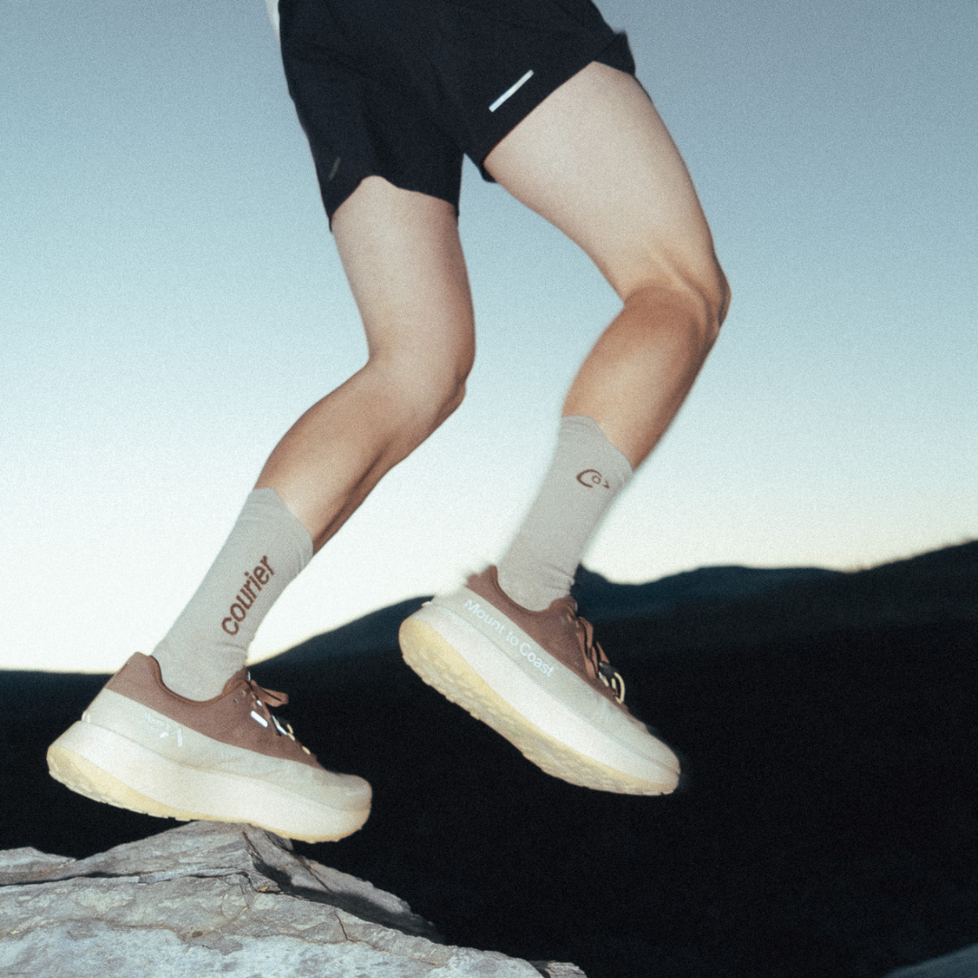 A person wearing beige and brown trail running shoes with matching socks jumps over a rock in an outdoor mountain setting at dusk.